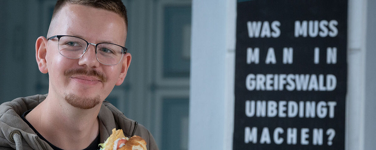 A student is holding a fish roll in his hand while looking very happy. Behind him is a sign saying "What is a must-do Greifswald?"