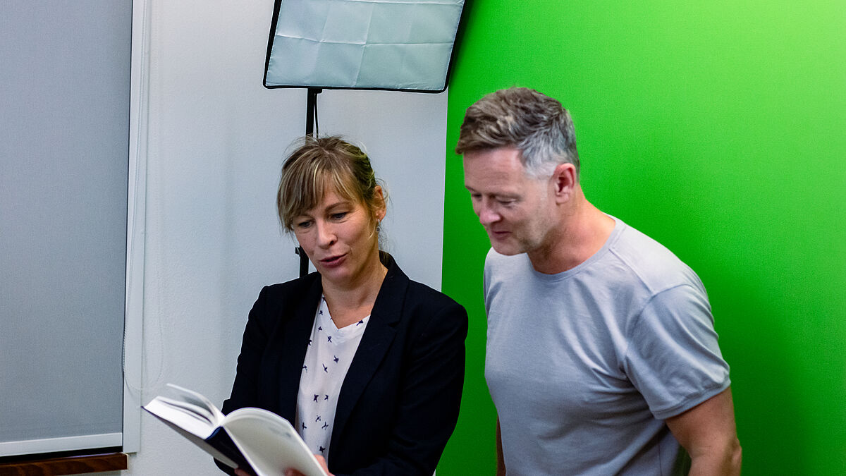 Zwei Personen stehen vor einem Greenscreen und lesen aus einem Buch vor.