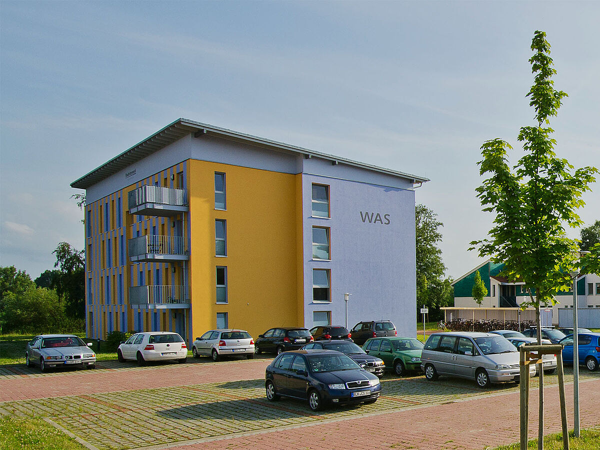 Modernes, rechteckiges Flachdachgebäude des Studentenwohnheim mit Parkplatz und Begrünung im Vordergrund.